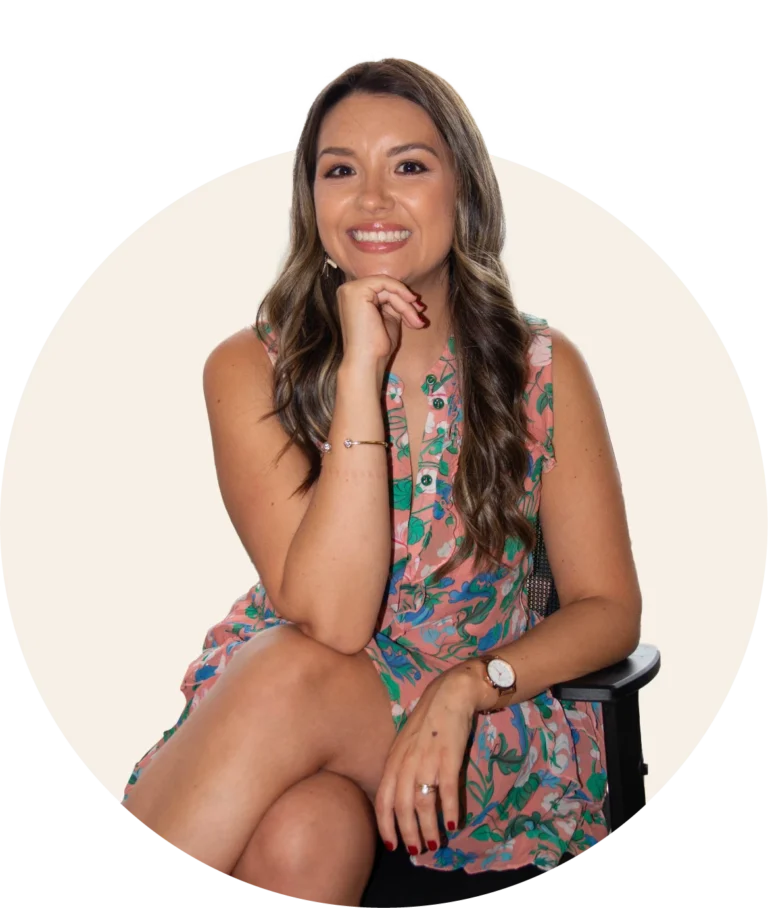 A professional headshot of Dr. Ana Maria smiling with long brown hair, wearing a floral patterned sleeveless dress. She is seated with her legs crossed and her hand resting under her chin in a circular frame.