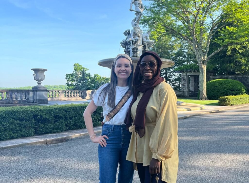 Two women, one in a white top and jeans, the other wearing a headscarf and a yellow tunic, stand smiling in front of a large stone fountain and manicured gardens on a sunny day.